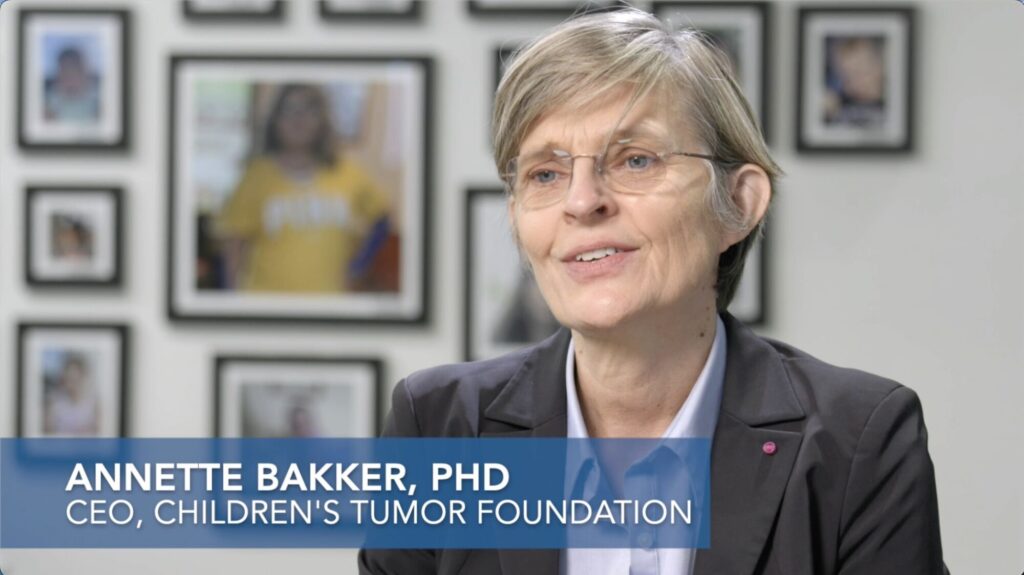 A woman with short hair and glasses is speaking, wearing a blazer and shirt. Text reads: "Annette Bakker, PhD CEO, Children's Tumor Foundation." Blurred photos are on the wall behind her.